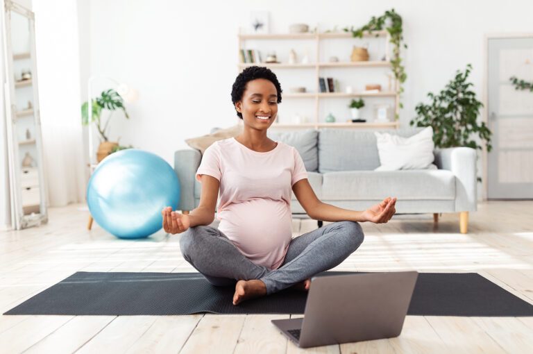 Belle femme enceinte méditant sur une vidéo en ligne sur un ordinateur portable à la maison. Jeune femme enceinte faisant du yoga pour atteindre l'équilibre intérieur, assise en posture de lotus dans son salon.